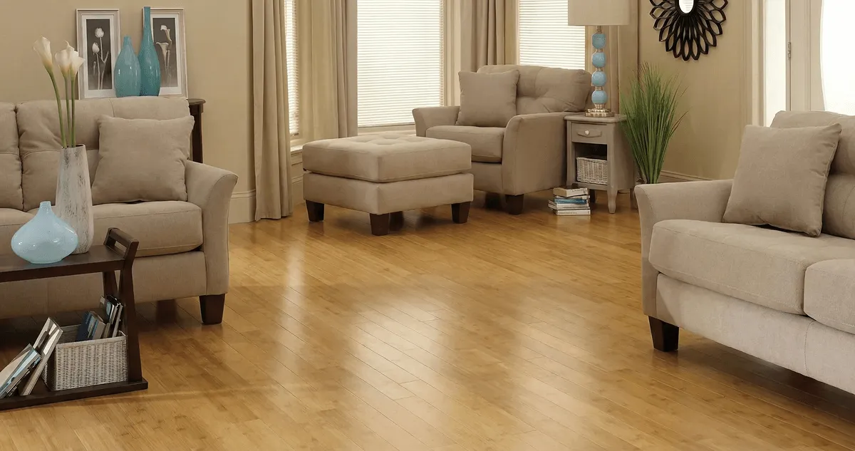 A living room with professionally installed bamboo flooring, beige sofas, a matching ottoman, a side table with a blue lamp, vases, framed pictures, and large windows with blinds and curtains in Evansville, IN